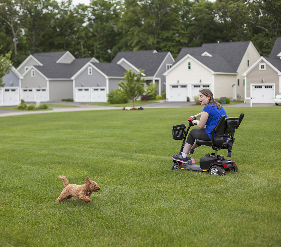 Young woman in wheelchair on grass with a small dog in front of affordable housing
